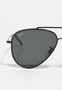 Černé kovové pilotní sluneční brýle s tmavě šedými skly. Obsahují výrazné logo Ray-Ban a tenký drátěný rám s nastavitelnými nosními podložkami.