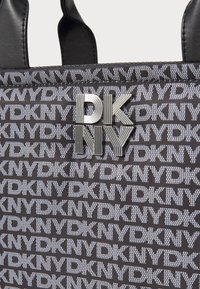 Saco tote em tecido preto com um padrão repetido do logótipo branco da DKNY e um emblema metálico prateado da marca na parte da frente. Textura suave.
