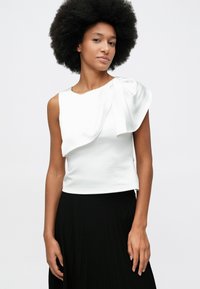 Woman with afro hairstyle wearing sleeveless white top with a large shoulder ruffle and a black pleated skirt against white background.