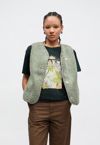 Zelená fleecová vesta s textúrovaným povrchom, zaobleným lemom a cvokmi, cez tmavé tričko s farebnou grafickou potlačou a hnedé nohavice.