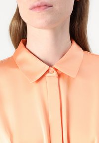 Blouse en satin pêche avec un col classique à bouton, une patte de boutonnage à l'avant et une texture lisse. Aucune motif ni embellissement visible.