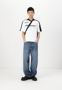 White polo shirt with black accents and logo, loose blue denim jeans, and chunky black shoes. Model stands with hands in pockets.