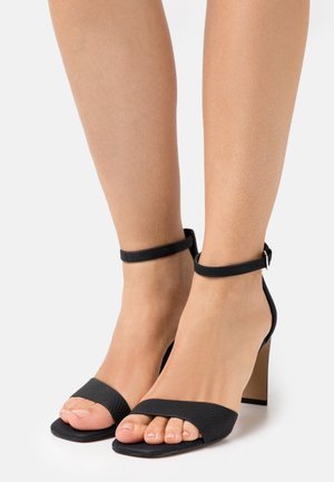 Fødder iført sorte højhælede sandaler med ankelstropper og strukturerede forreste stropper på en simpel hvid baggrund.