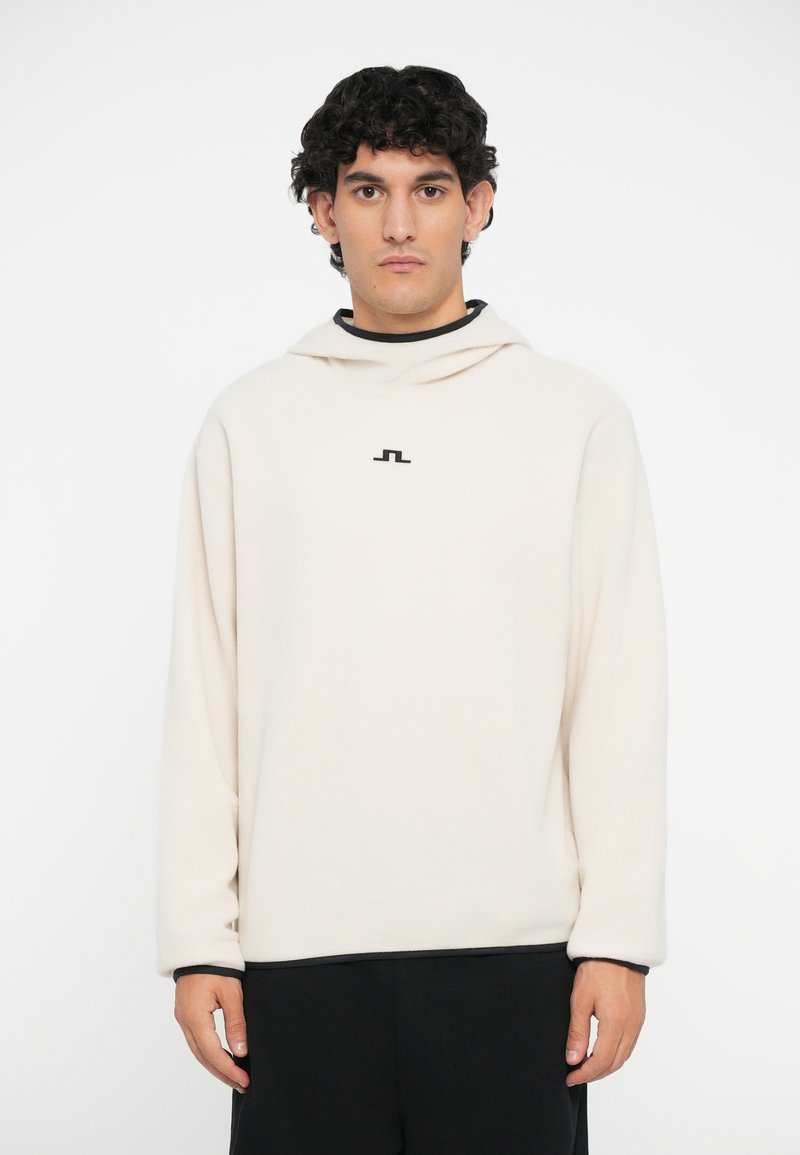 Cremefarbener Hoodie mit lockerer Passform, der ein schwarzes Logo auf der Brust, einen hohen Kragen und lange Ärmel hat. Weicher Stoff mit feinen Nähten.