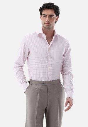 Mann trägt ein hellrosa kariertes Hemd, in graue hoch taillierte Faltenhose gesteckt, steht mit einer Hand in der Tasche.