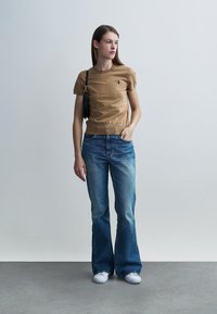 Κοντομάνικη μπλούζα σε χρώμα ταμπά με λογότυπο, συνδυασμένη με ανοιχτόχρωμο μπλε τζιν καμπάνα και λευκά αθλητικά παπούτσια. Κρατάει μια μικρή μαύρη τσάντα.