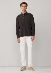 Polo a maniche lunghe di colore marrone scuro con colletto a tre bottoni, abbinato a pantaloni bianchi e scarpe beige. Tessuto morbido, vestibilità casual.