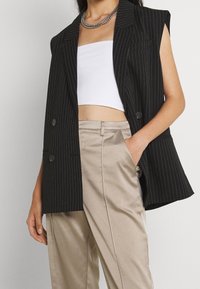 Svart randig dubbelknäppt väst utan ärmar, tillsammans med en vit crop top och beiga satinbyxor. Har knappar och sidfickor.