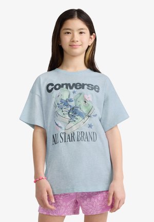 Fille portant un t-shirt Converse bleu clair avec un motif de baskets et un short rose à motifs, debout sur un fond blanc.