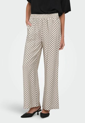 Beige bukser med vide ben og sorte polkaprikker, elastisk talje, modellens venstre hånd i lommen, kombineret med sorte slingback-hæle og sort top.