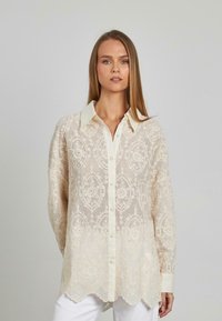 Spitzenbluse in Creme mit gestickten Blumenmustern, knöpfbarem Vorderteil und spitzem Kragen. Verfügt über lange Ärmel und einen welligen Saum.