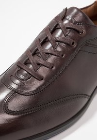 Chaussure en cuir marron à bout arrondi, avec surface texturée, couture décorative et six œillets pour les lacets. Finition lisse et design élégant.