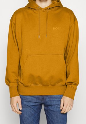 Persoon draagt een mosterdgele hoodie met een zak aan de voorkant en een subtiel "BOSS"-logo op de borst, gecombineerd met een blauwe spijkerbroek.