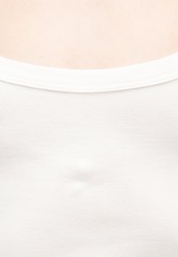 Κοντινό πλάνο ενός λευκού τοπ χωρίς μανίκια που δείχνει το λαιμόκοψη και την περιοχή πάνω από το στήθος, πάνω σε ανοιχτό φόντο.