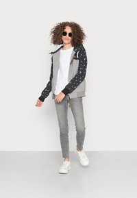 Casaco cinza com fecho éclair e mangas pretas estampadas, combinado com uma t-shirt branca, jeans cinza e ténis brancos. Cabelo encaracolado e óculos de sol.
