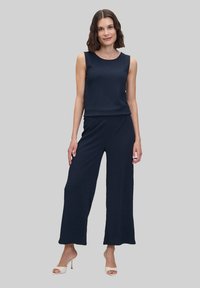 Donkerblauwe mouwloze top gecombineerd met bijpassende wijde broek. Gladde, gestructureerde stof. Model draagt neutrale hoge hakken.