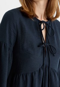 Blusa azul marino hecha de tejido texturizado, con un cierre de lazo en el escote y mangas abullonadas, con un diseño suelto y en capas.