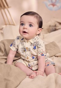 Īsroku bērnu oglīte krēmkrāsā ar krāsainiem grafiskajiem attēliem. Aprīkota ar pogām un ērtu piegriezumu. Novietota uz mīksta, gaiša auduma fona.