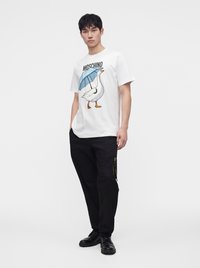 Νέος άνδρας με λευκό μπλουζάκι Moschino με γραφικό πάπιας που κρατάει μπλε ομπρέλα, μαύρο παντελόνι και μαύρα παπούτσια, στέκεται σε απλό φόντο.