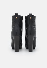 Botines negros con acabado de cuero liso, tacones altos y gruesos, y paneles decorativos con patrones en la parte trasera. Cierre de cremallera en los laterales.