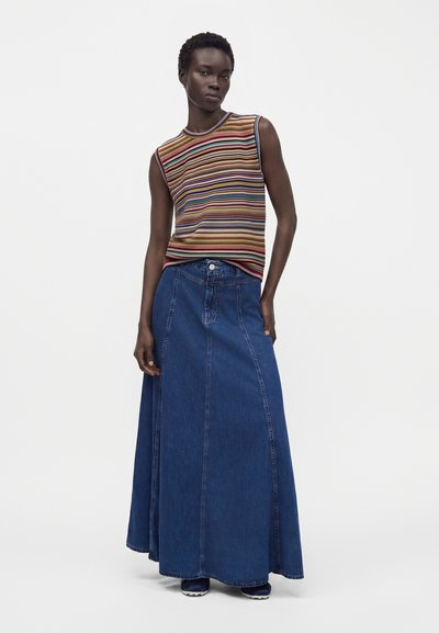 Personne portant un haut sans manches à rayures multicolores et une longue jupe en jean bleue taille haute, debout devant un fond uni.