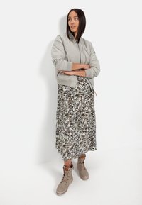 Hellgraue Bomberjacke mit gerippten Bündchen, kombiniert mit einem floralen Midi-Kleid in Grün- und Brauntönen, dazu beiges Schnürstiefel.