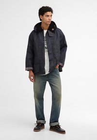 Casaco impermeável azul-marinho com design de gola e botões, duas bolsos frontais e interior xadrez. Combinado com jeans de corte relaxado e sapatos castanhos.