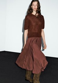 Maglione corto a maniche corte in maglia pelosa marrone, abbinato a una gonna marrone fluente di lunghezza media e stivali al ginocchio in suede marrone su uno sfondo scuro.