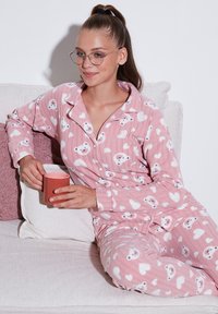 Set de pijamale roz dintr-un material moale, cu modele de urși albi și inimioare, cu o bluză cu nasturi și mâneci lungi, purtat de o persoană care ține o cană.