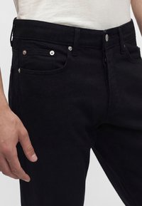 Primo piano di una persona che indossa jeans neri in denim con rivetti e bottone argentati, abbinati a una camicia beige chiaro infilata dentro.