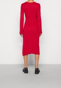 Robe rouge moulante en tricot, manches longues, texture côtelée, longueur mi-mollet, fente latérale, associée à des chaussures noires épaisses.