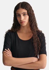 Sort t-shirt med korte ærmer og rund hals, glat tekstur, tætsiddende design, båret med armene korslagt. Baggrunden er ensfarvet hvid.