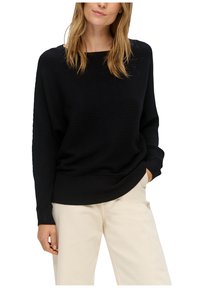 Schwarzer gerippter Pullover mit weitem Ausschnitt, fallenden Schultern und langen Ärmeln. Kombiniert mit hellbeigen Hosen, die eine lockere Passform betonen.