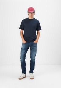 Marine t-shirt, blå jeans med let slid, hvide sneakers med brune såler, og en rød kasket, båret af en mandlig model der står.