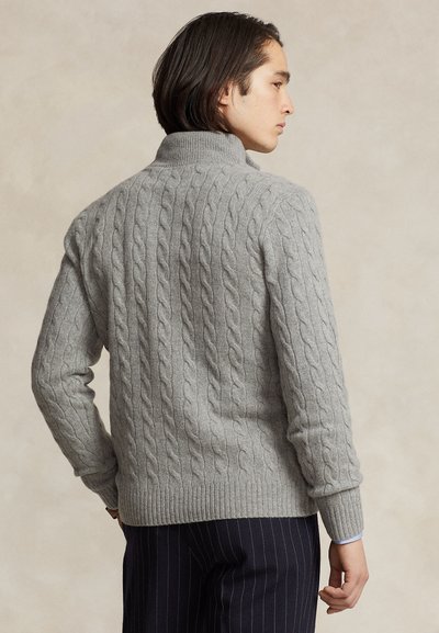Uomo visto di profilo indossa un maglione a trecce grigio chiaro con collo alto e pantaloni scuri a righe sottili su uno sfondo neutro.