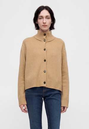 Kvinde iført en beige knappet cardigan med høj krave og blå jeans, stående mod en ensfarvet hvid baggrund.