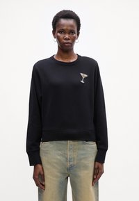 Sudadera negra con cuello y puños de canalé, que presenta un diseño de copa de martini adornada con cristales en el lado izquierdo del pecho. Llevada con jeans de color azul claro.