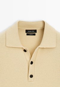 Polo shirt di lana color crema con colletto a costine, tre bottoni neri e una finitura a maglia testurizzata. Etichetta del marchio visibile all'interno del colletto.