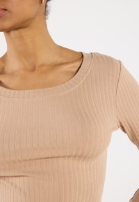 Langermet topp i beige ribbestrikket stoff, med bred scoop-hals og tettsittende silhuett, som fremhever vertikale teksturdetaljer.