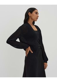 Cardigan noir en tricot avec un design court, à l'avant ouvert, et des manches longues mettant en valeur un motif texturé et tissé.