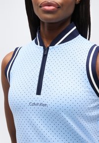 Κοντινό πλάνο γυναίκας που φοράει αμάνικο, ανοιχτό μπλε, πουά τοπ Calvin Klein με φερμουάρ, με σκούρο μπλε λεπτομέρειες στο γιακά και τα μανίκια.