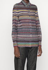 En långärmad skjorta med knappar med färgglada zigzag-ränder som bärs med svarta byxor, visad från mitten av nacken till mitten av låren på en mörkhyad person.