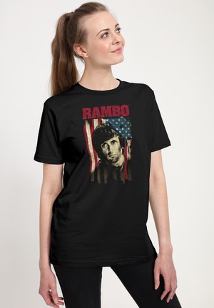 Kvinde med langt brunt hår i hestehale iført en sort T-shirt med et Rambo-motiv foran en baggrund med det amerikanske flag.