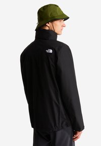 Mies, jolla on oliivinvihreä kalastajanhattu ja musta North Face -takki, nähtynä takaa ja katsomassa sivulle valkoista taustaa vasten.