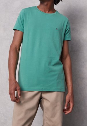 T-shirt vert à manches courtes en coton avec un col rond et un petit logo sur la poitrine. Associé à un pantalon beige. Texture lisse.