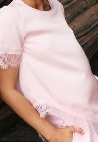 Personne enceinte portant une robe rose pâle avec un bord en dentelle aux manches et à l'ourlet, main reposant dans la poche sur fond sombre.