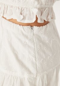 Tissu blanc brodé avec des motifs floraux et un col froncé. Dispose d'une fermeture éclair discrète à l'arrière. Texture douce.