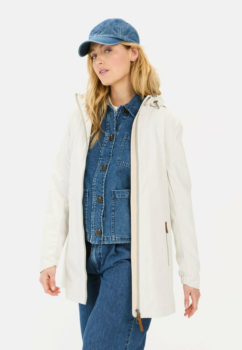 Witte lichtgewicht jas met een capuchon, ritssluiting en zijzakken, gedragen over een blauwe spijkeroutfit, afgewerkt met een blauwe spijkermuts.