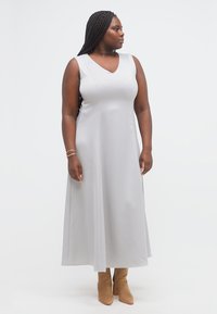 Γυναίκα με μακριά πλεξούδα, φοράει ένα αμάνικο μακρύ γκρι φόρεμα και ταμπά μποτάκια μέχρι τον αστράγαλο, κοιτάζοντας προς τα αριστερά της.