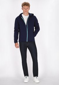 Felpa con zip color navy, con una cerniera azzurro chiaro, indossata sopra una t-shirt bianca e abbinata a jeans scuri e scarpe da ginnastica bianche. Texture liscia, design casual.